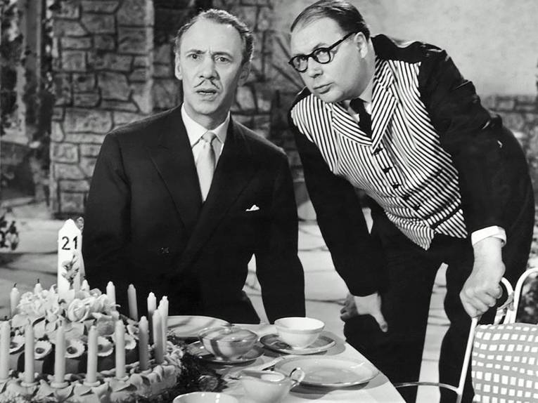 Zu sehen ist ein Mann im Anzug, der an einem gedeckten Kaffeetisch sitzt, auf dem ein Kuchen mit vielen Kerzen steht. Er macht einen verwirrten Eindruck. Ein anderer Mann in einer gestreiften Weste beugt sich zu ihm herunter.